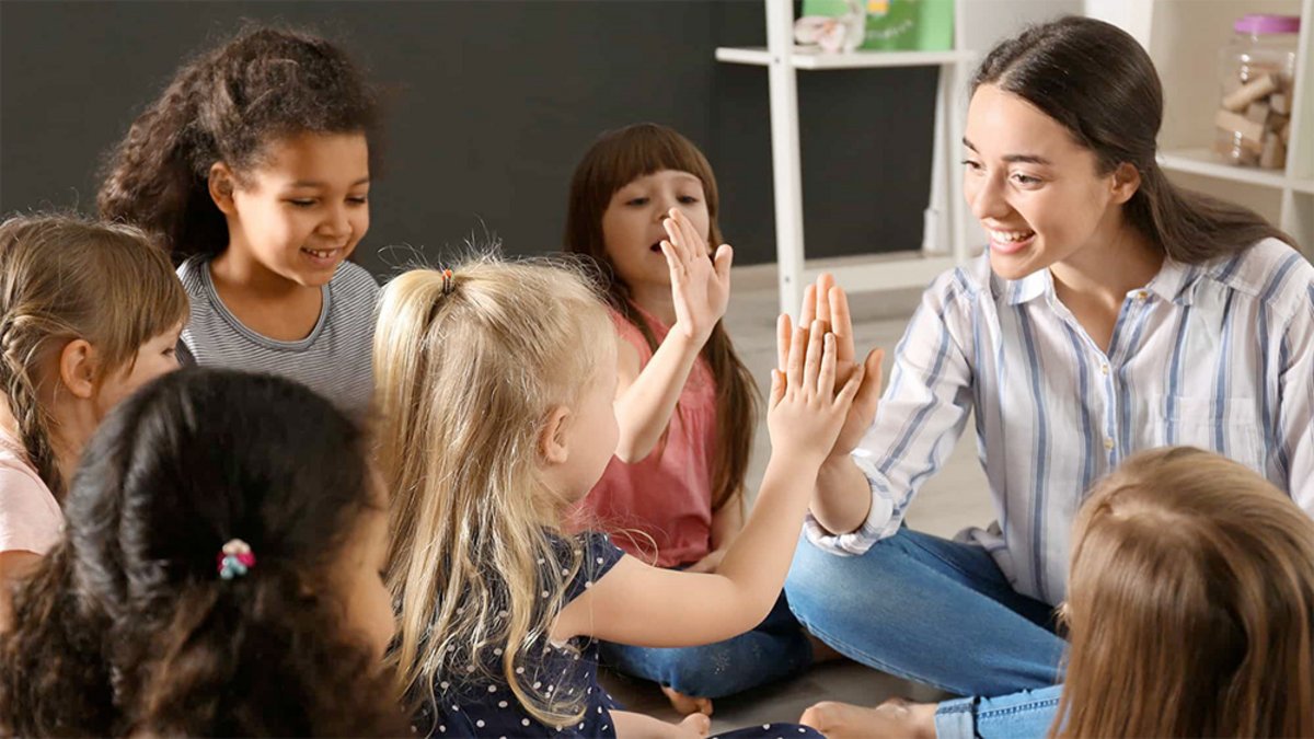 Das Foto zeigt eine junge Frau und sechs Kindergartenkinder, die auf dem Fußboden im Kreis sitzen. Die junge Frau ist in Interaktion mit den Kindern und spielt ein Klatschspiel.