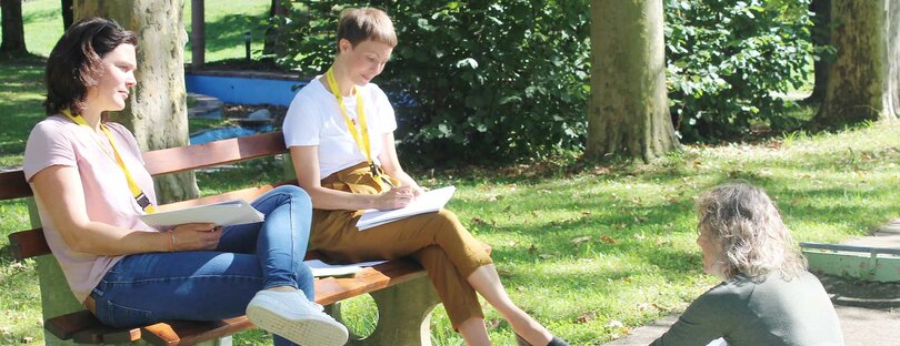 Das Foto zeigt drei Personen in einem Park. Zwei sitzend auf einer Bank, eine dritte Person den auf der Bank gegenüber auf dem Boden sitzend. Die Personen sind mit Arbeitsmaterialien in den Händen in ein Gespräch vertieft