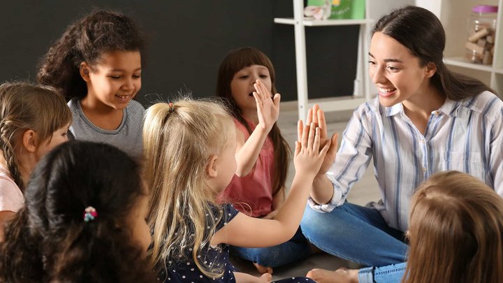 Das Foto zeigt eine junge Frau und sechs Kindergartenkinder, die auf dem Fußboden im Kreis sitzen. Die junge Frau ist in Interaktion mit den Kindern und spielt ein Klatschspiel.